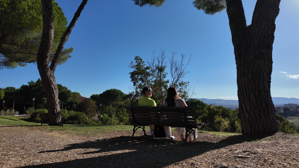 Casal sentado em banco de madeira sob pinheiros contemplando a paisagem das colinas da Toscana em dia ensolarado
